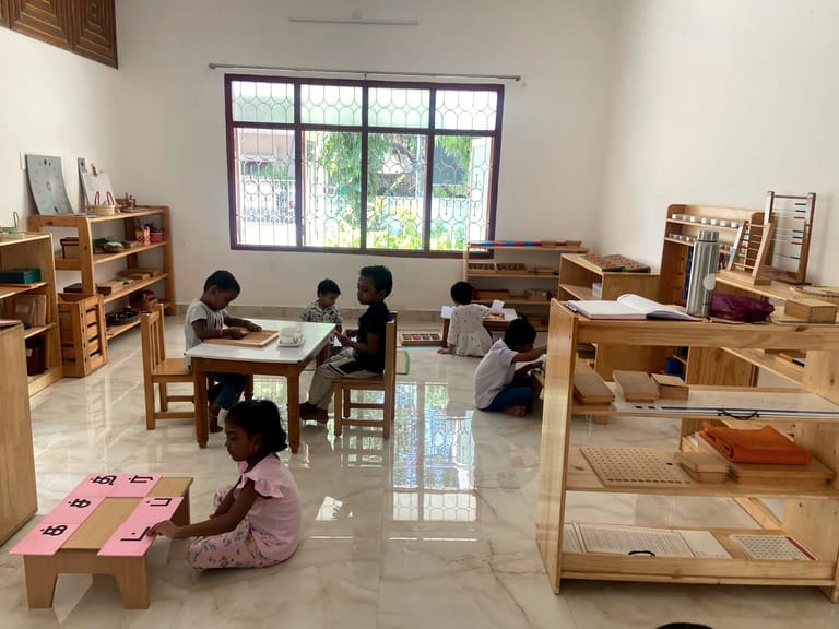 Children working at low tables and on mats beside a large classroom window.