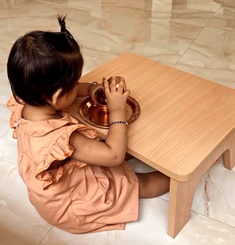 A child polishes a small brass vessel at a low wooden table.