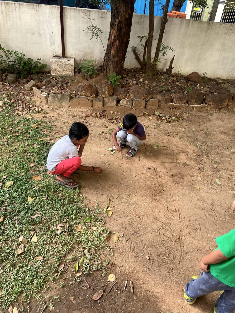 Two children crouch beneath a tree and look closely at the bare ground.