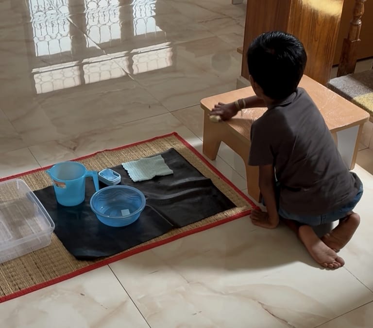 A child washes a small wooden table beside a basin, jug and cloths.