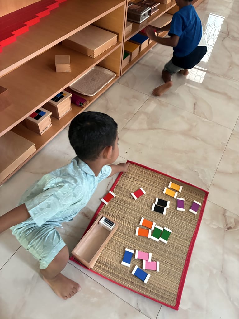 A child pairs coloured tablets on a floor mat beside the classroom shelves.